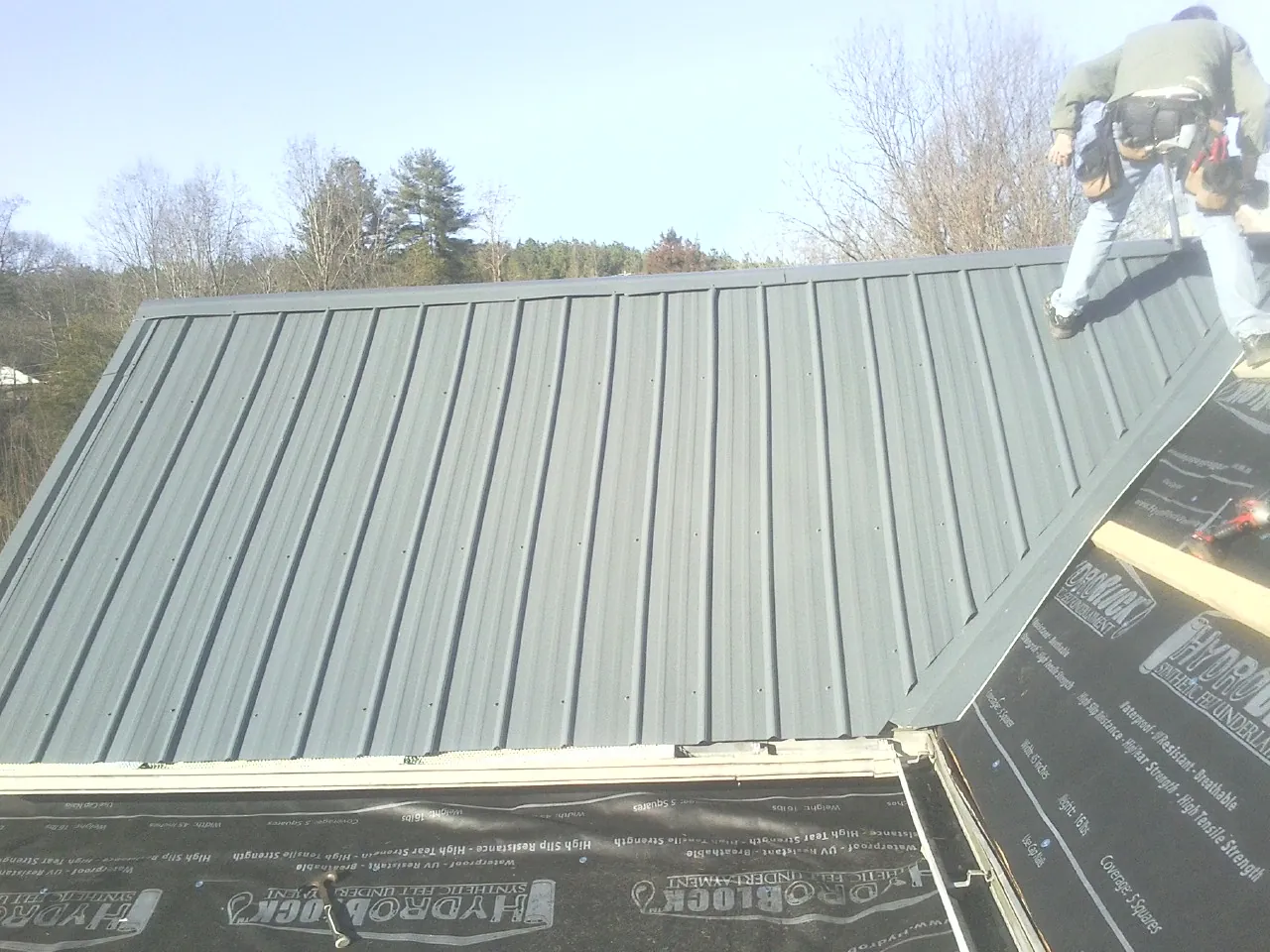 Crew installing dark green standing seam panels over synthetic underlayment in Maple City