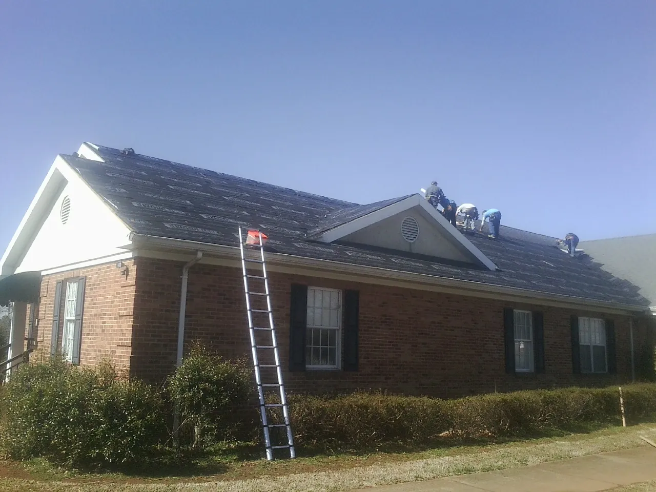 Crew tearing shingles off a brick ranch roof with a ladder set — Emergency Roof Repair work by our crew in Maple City