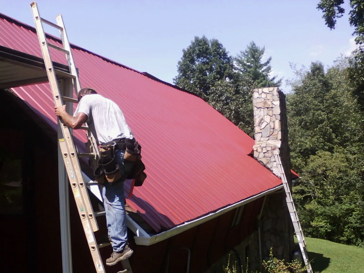 Roofer on a ladder beside a red metal roof and stone chimney — Soffit & Fascia Repair by Roofing Brothers in Maple City